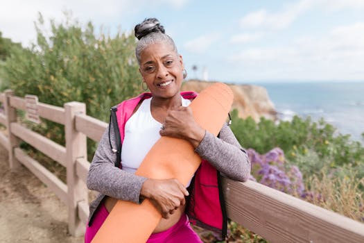 Smiling senior woman holding a yoga mat by the coastline, enjoying wellness and nature.