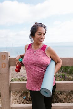 Smiling senior woman holding yoga mat outdoors by the beach, embracing active lifestyle.
