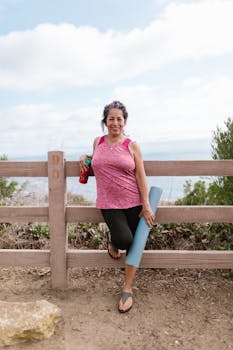 A senior woman smiling while holding a yoga mat and thermos by a wooden fence overlooking the ocean.