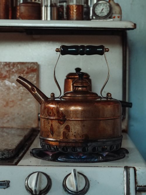 Free A weathered copper kettle heating on a vintage gas stove, exuding rustic charm. Stock Photo