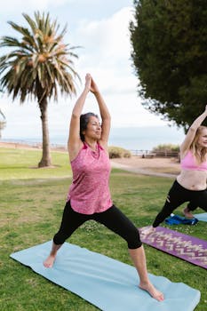 Two women performing yoga outdoors on mats near a beach, promoting fitness and well-being.