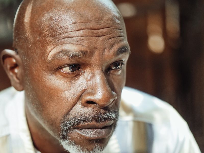 A close-up portrait of a mature Black man, bald with a short grey goatee, looking off to the side with a calm, thoughtful expression in warm light.