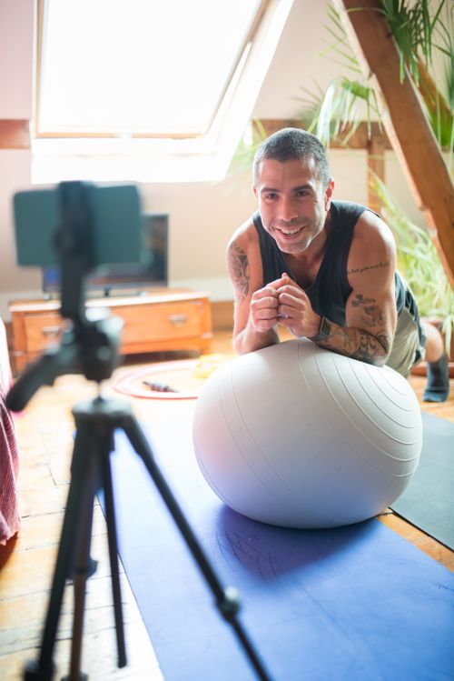 Free Smiling man exercises on a yoga mat with an exercise ball, capturing workout on video indoors. Stock Photo