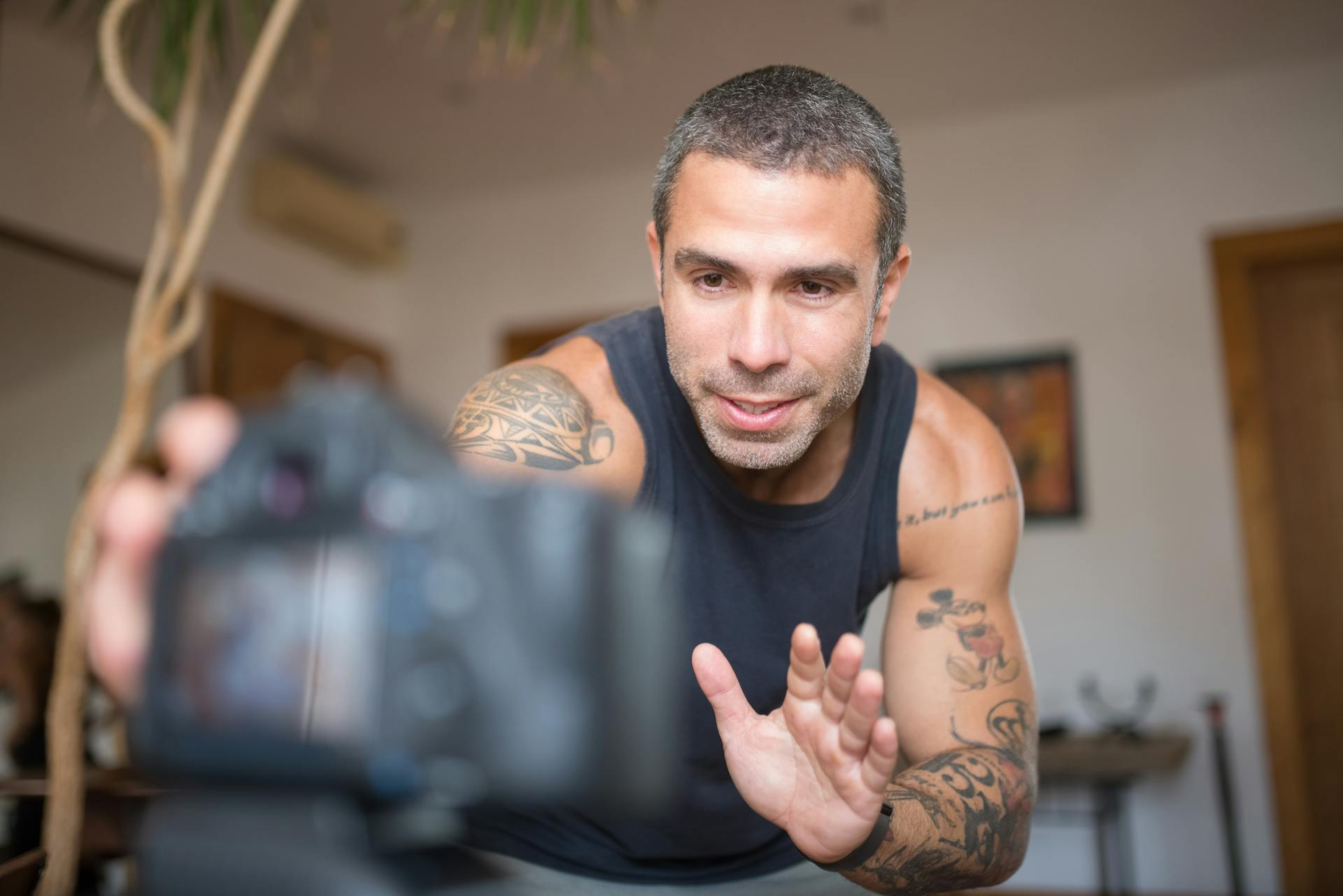 Tattooed man in a tank top recording a fitness video at home, focusing on training.