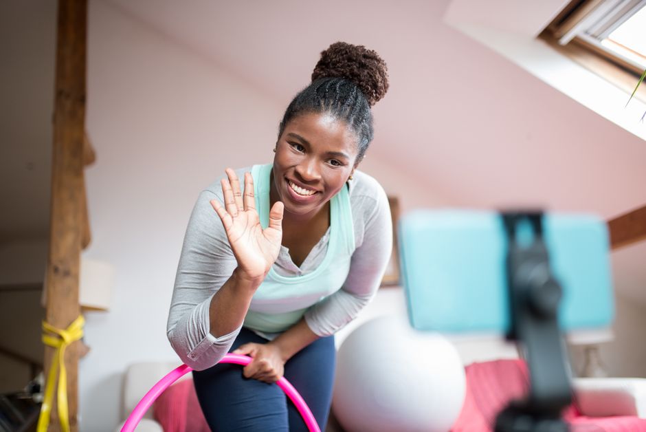 A smiling woman waves to the camera while recording a fitness vlog indoors.