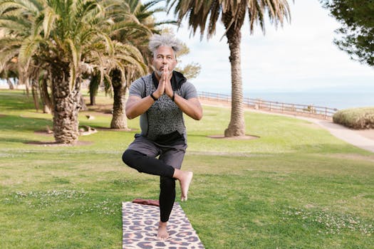 A man performs a tree pose on a yoga mat in a sunny park, surrounded by palm trees.