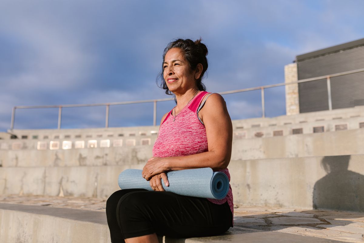 A smiling woman sitting outdoors with her yoga mat