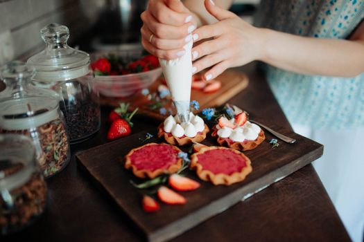 Crop unrecognizable female confectioner putting cream on colorful tasty cookies with berries on wooden board in kitchen