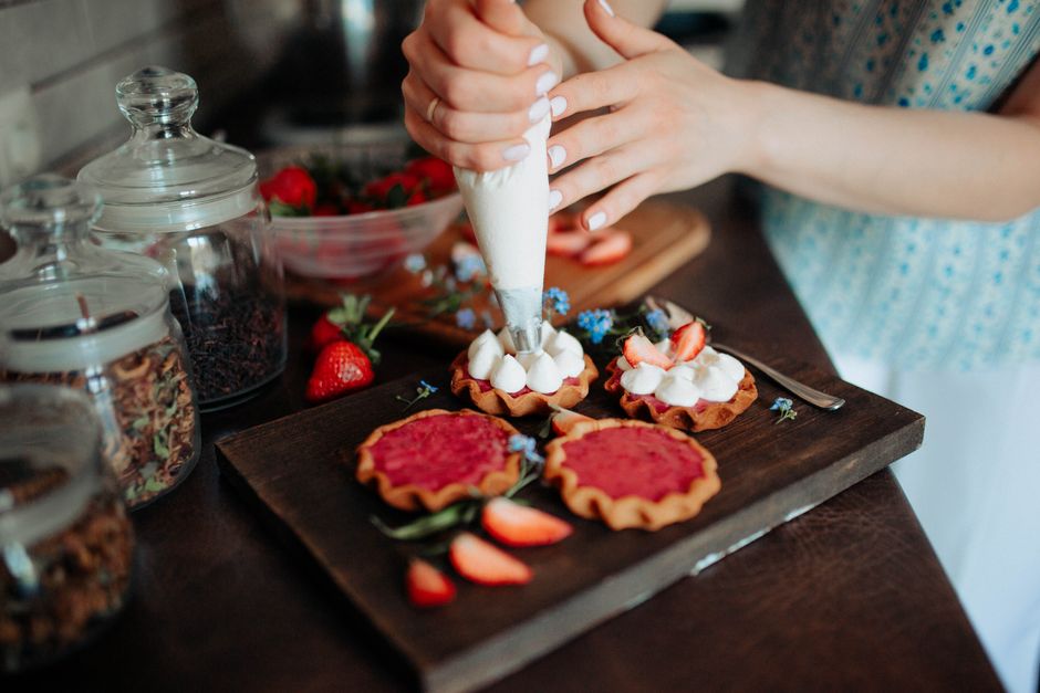 Crop unrecognizable female confectioner putting cream on colorful tasty cookies with berries on wooden board in kitchen
