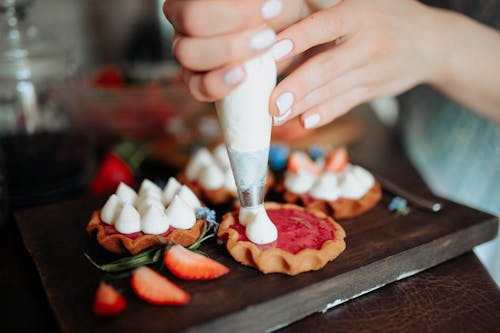 Crop anonymous female adding cream on homemade cookies with strawberries placed on wooden board in kitchen in daylight