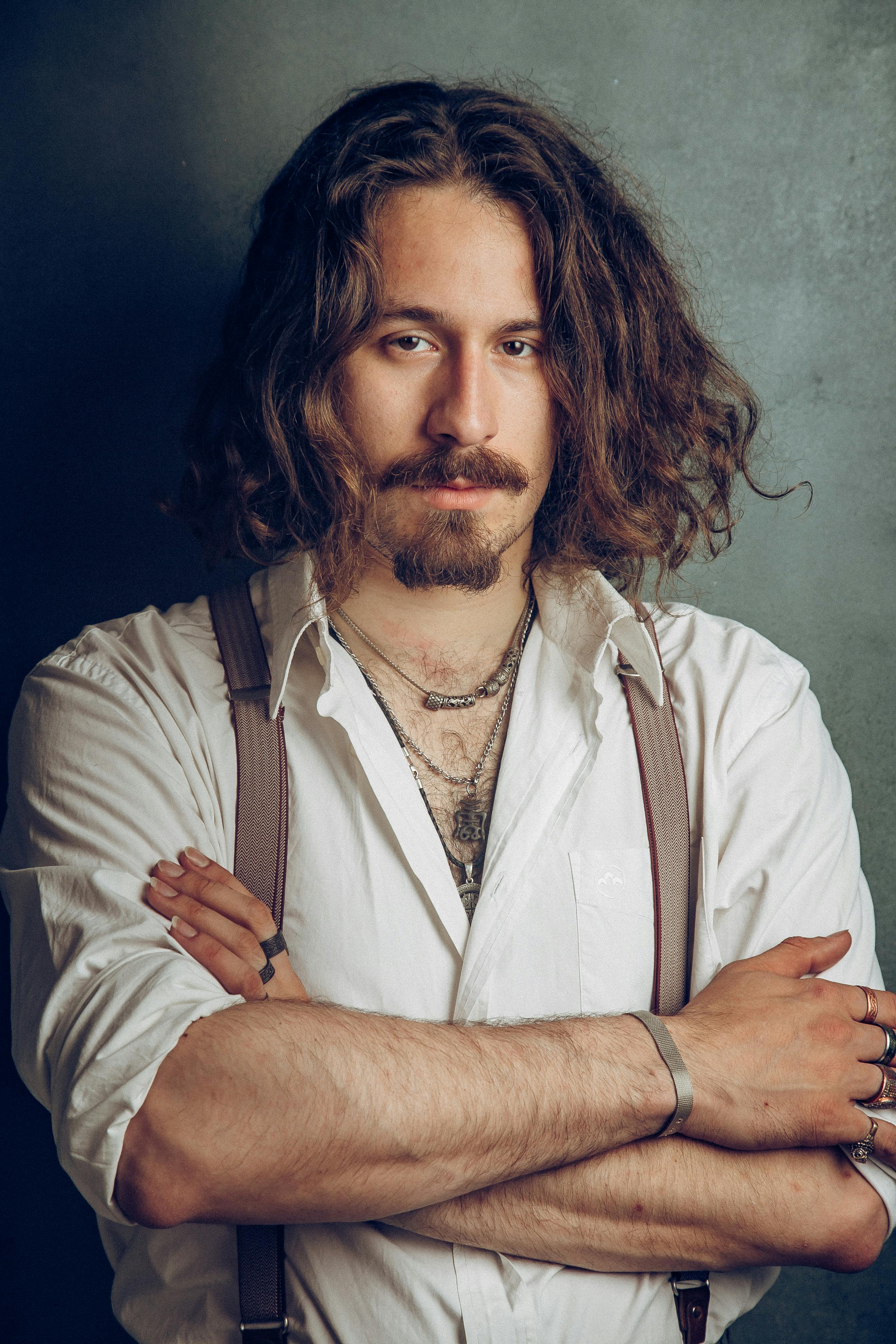 Studio portrait of a man with long hair, beard, and suspenders, arms crossed.