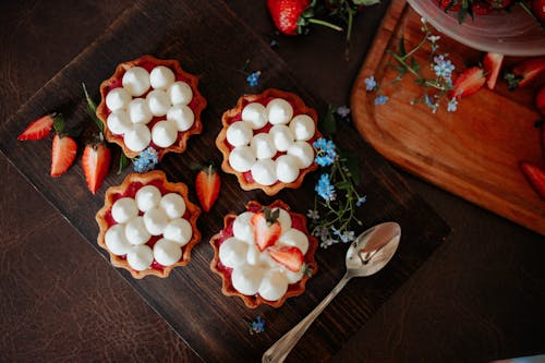 From above of delicious cookies decorated with cream and strawberries placed on wooden board with spoon and flowers on table in kitchen