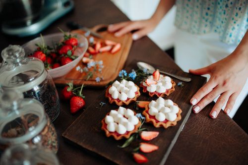 Free Crop anonymous female standing near table with delicious cookies decorated with cream and strawberries placed on wooden board in kitchen Stock Photo