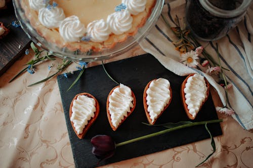 Top view of delicious homemade desserts decorated with cream placed on table near cake and flowers in kitchen