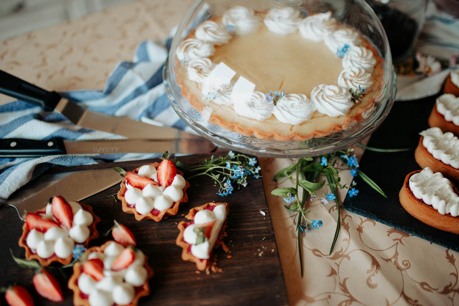 Pie decorated with cream on tray with glass cap near flowers and different desserts on cutting boards near knives on table in bright kitchen