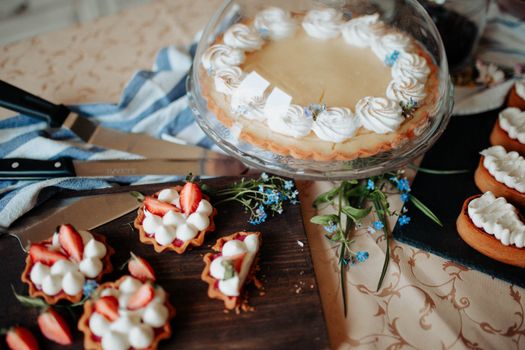 Pie decorated with cream on tray with glass cap near flowers and different desserts on cutting boards near knives on table in bright kitchen