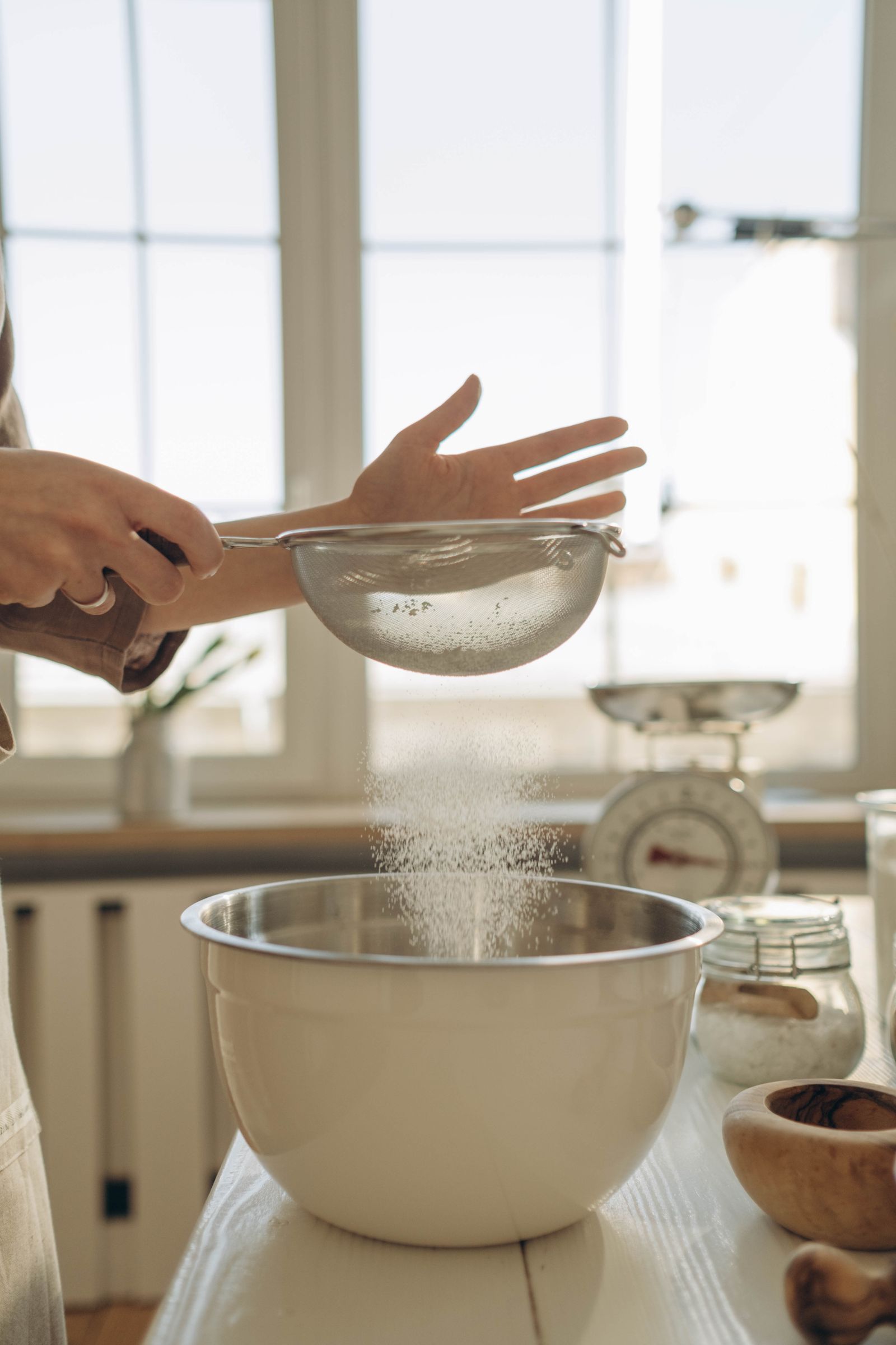Hands sifting white gluten-free flour into a mixing bowl on a sunlit kitchen counter