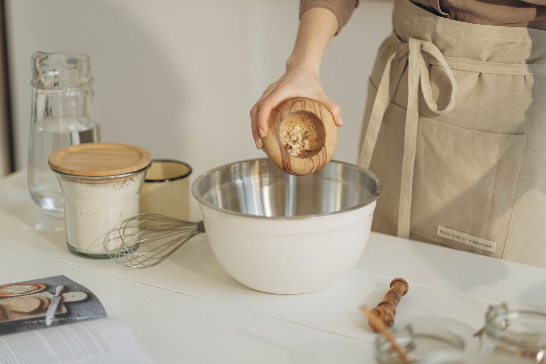 A woman pours ingredients into a mixing bowl, surrounded by baking tools in a cozy kitchen.