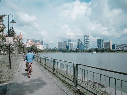 Free A cyclist rides along a waterfront path with a view of a modern city skyline under a cloudy sky. Stock Photo