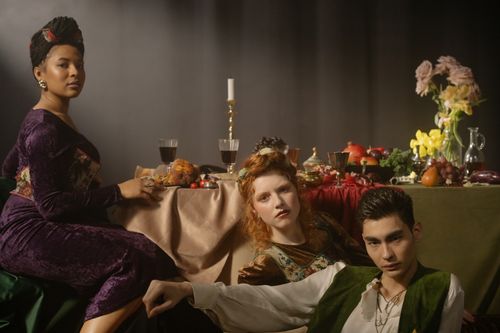 Free Three elegantly dressed individuals in a Renaissance-inspired setting with food and wine on a table. Stock Photo