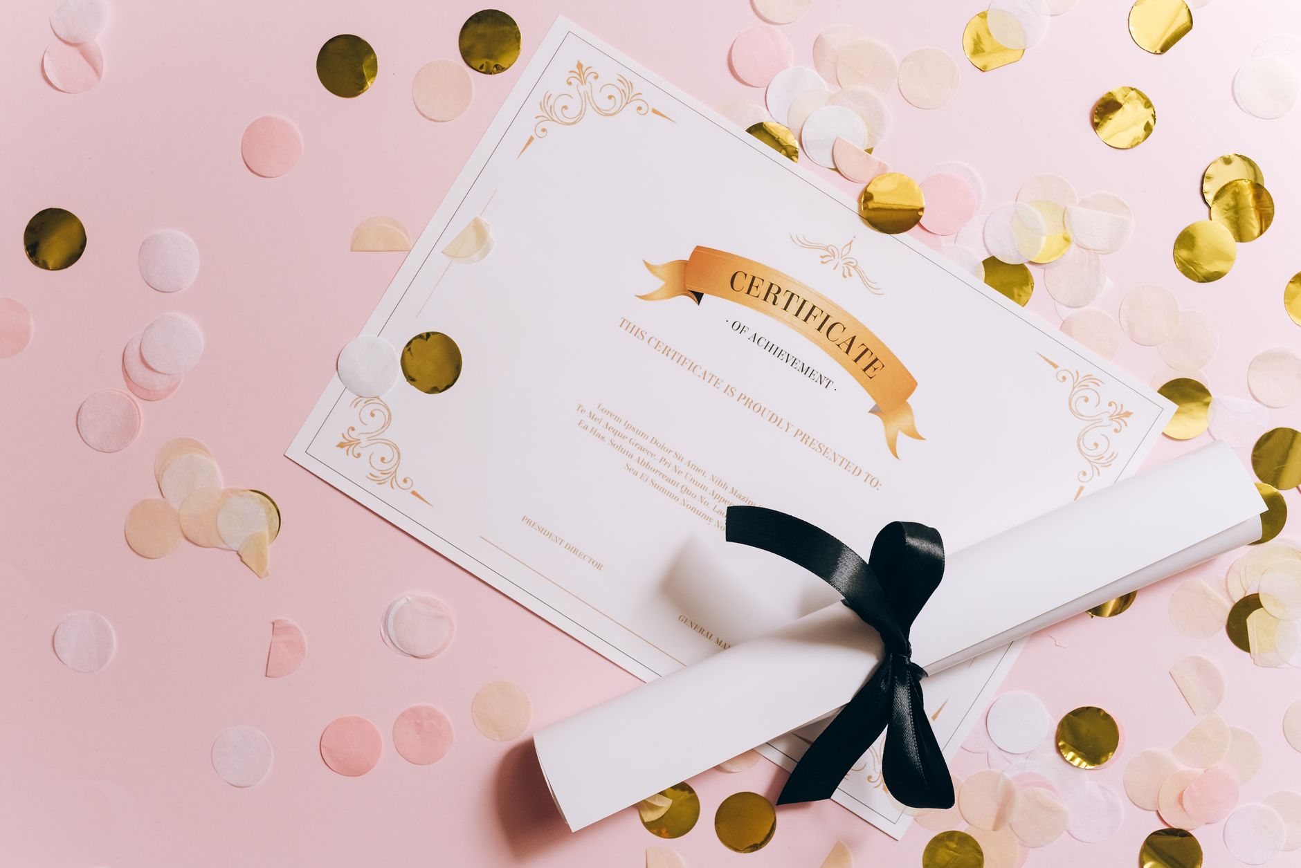 Certificate of achievement and diploma roll with ribbon on a pink confetti background.