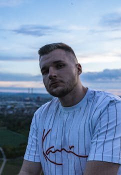 Portrait of a bearded man in a striped shirt during sunset, with a cityscape and clouds in the background.