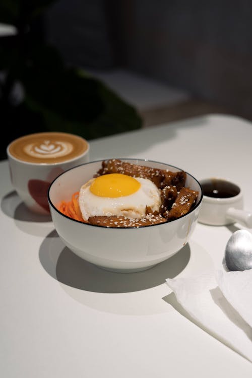 A Bowl of Noodles with Egg and Meat Beside the Cup of Cappuccino