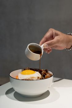 Hand pouring sauce over a bowl with egg and meat on a white table.