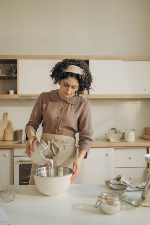 Free Woman in a cozy kitchen pouring ingredients into a bowl, wearing an apron and brown blouse. Stock Photo