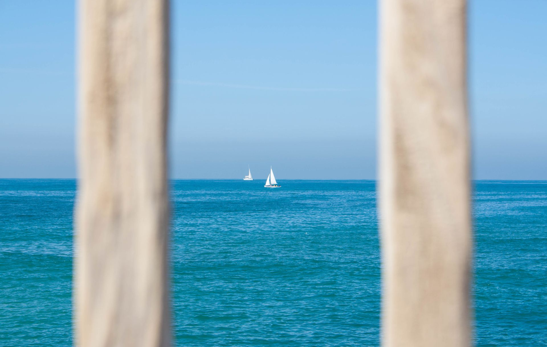A tranquil seascape with sailboats viewed through wooden posts on a sunny day.