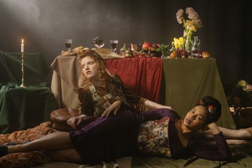 Free Two women in a Renaissance-style setup with fruits, flowers, and a candlelit table. Stock Photo