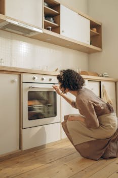 A woman kneeling to check the oven temperature in a cozy kitchen, wearing a brown dress and apron.