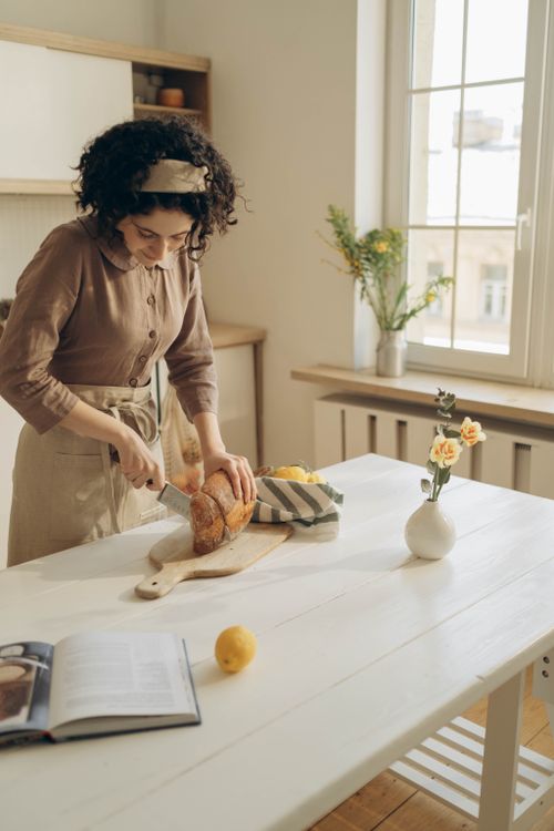 Free A woman slices fresh bread on a wooden chopping board in a bright kitchen setting. Stock Photo