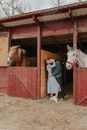 Side view of trendy man kissing and embracing girlfriend between stallion muzzles in countryside stall