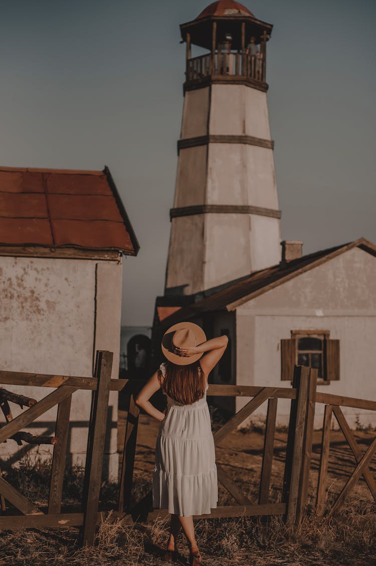 Back View Of A Woman Wearing Dress And Hat Looking At Old Building With Observation Tower