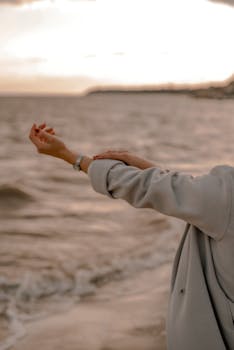 A close-up of a woman's hand with a watch, reaching towards the sea at sunset.