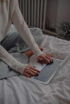 A woman with painted nails sitting on bed, using a laptop in a comfortable indoor space.