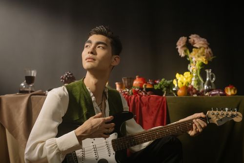 Free A young Asian man with guitar in an artistic setup featuring fruits and flowers. Stock Photo