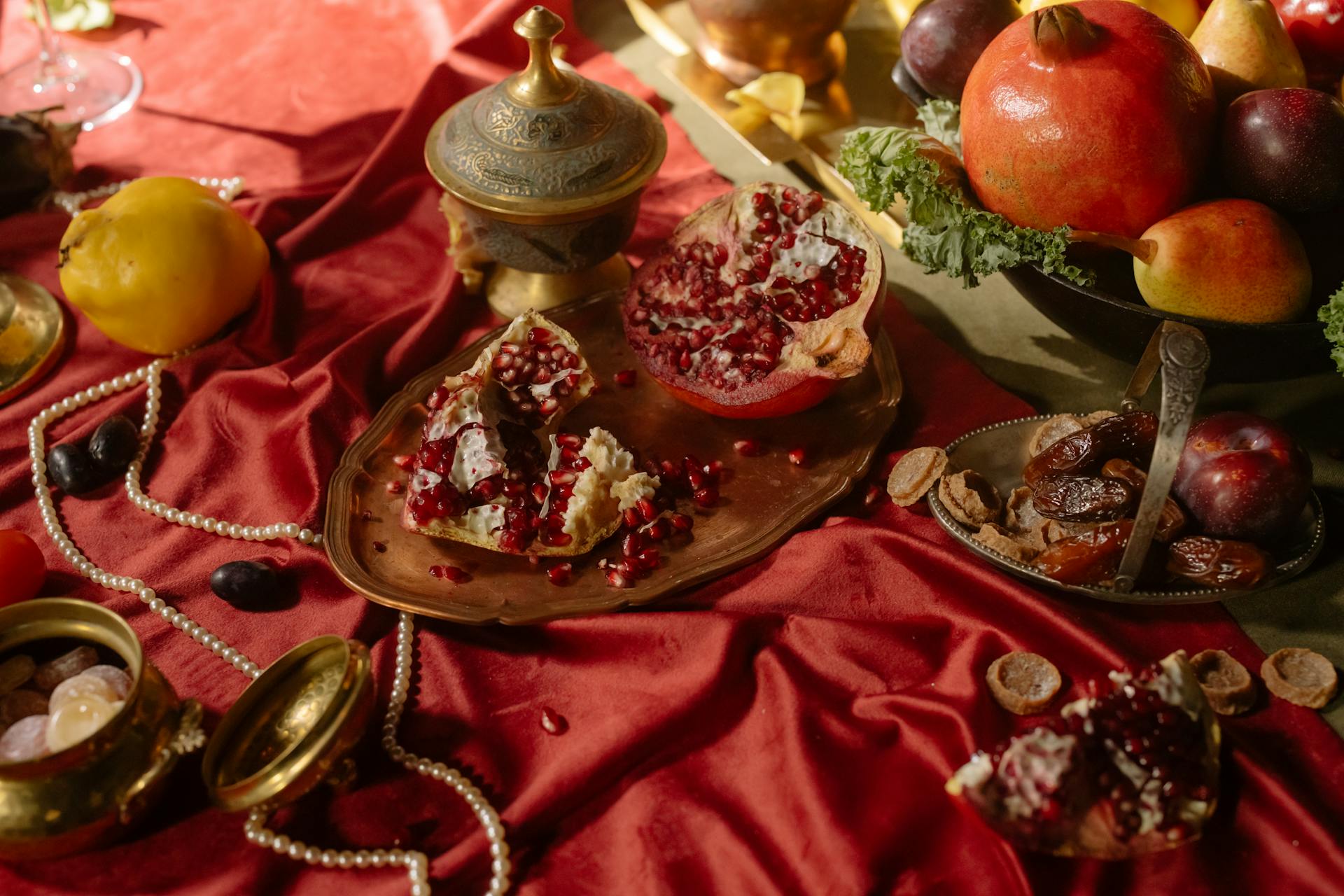 Luxurious still life featuring fruits, pearls, and a brass tray on a rich red cloth.