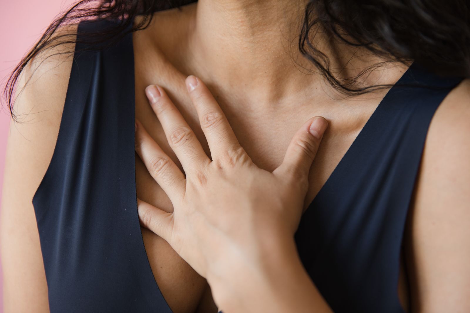 A woman in a sleeveless blue top resting one hand flat against the center of her chest, the spot where reflux is felt as heartburn