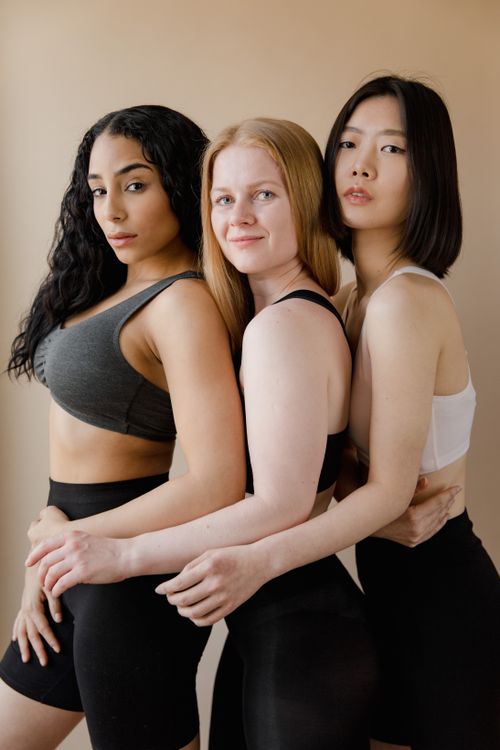 Free Three diverse women in sports bras embracing in a warm, minimalist setting. Stock Photo