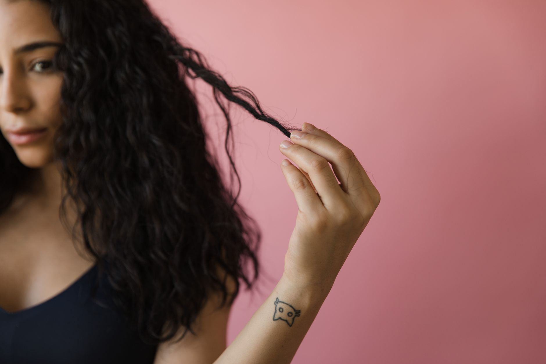 A woman with tattooed wrist examines her curly hair against a pink background.