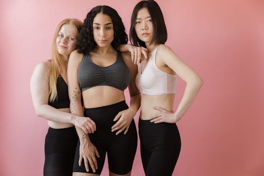 Three diverse women posing in activewear against a pink background, embracing fitness and body positivity.