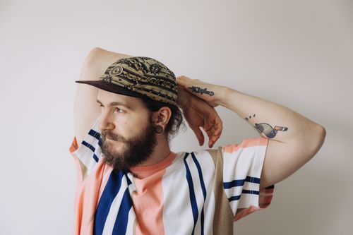Free Stylish man posing with a beard, cap, and colorful tattoos on a minimalist background. Stock Photo