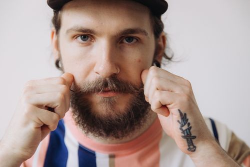 Free Detailed close-up of a young, bearded man with a nose piercing and tattoo pulling his mustache. Stock Photo