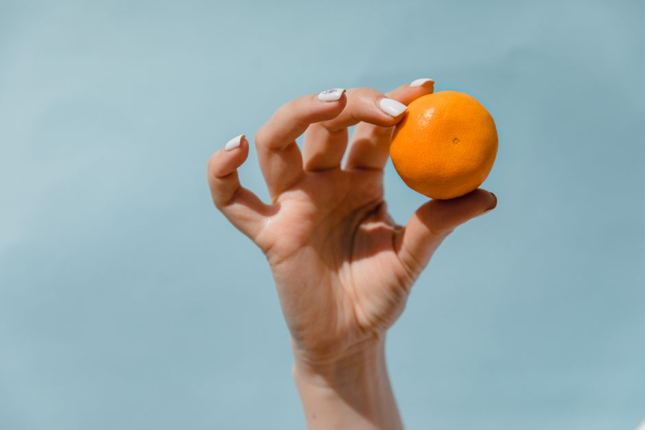 A hand with manicured nails elegantly holding a juicy fresh mandarin against a blue background.