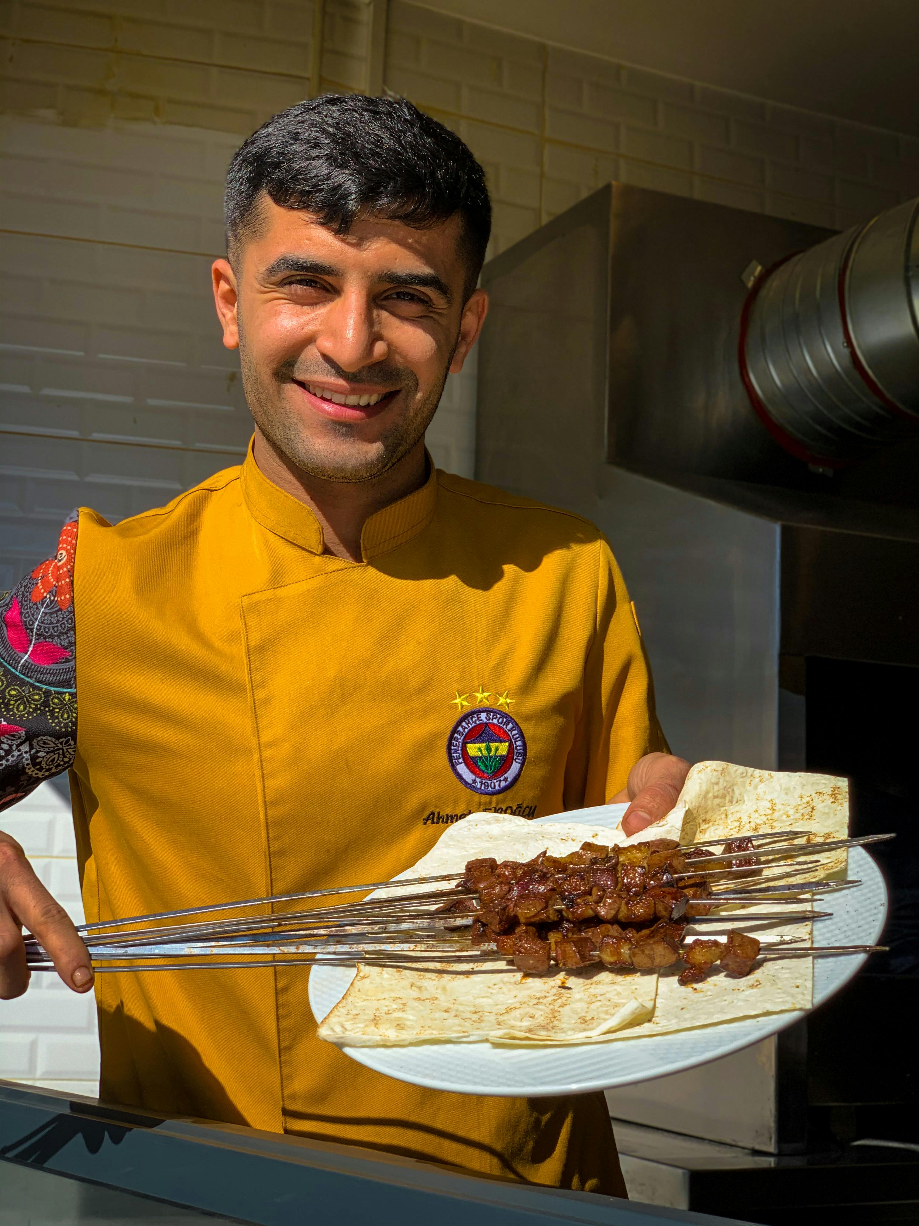 Smiling chef in yellow uniform presenting mouthwatering grilled meat skewers indoors.