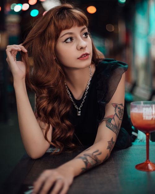 Free Young woman with tattoos posing at a bar counter with a cocktail, looking sideways. Stock Photo