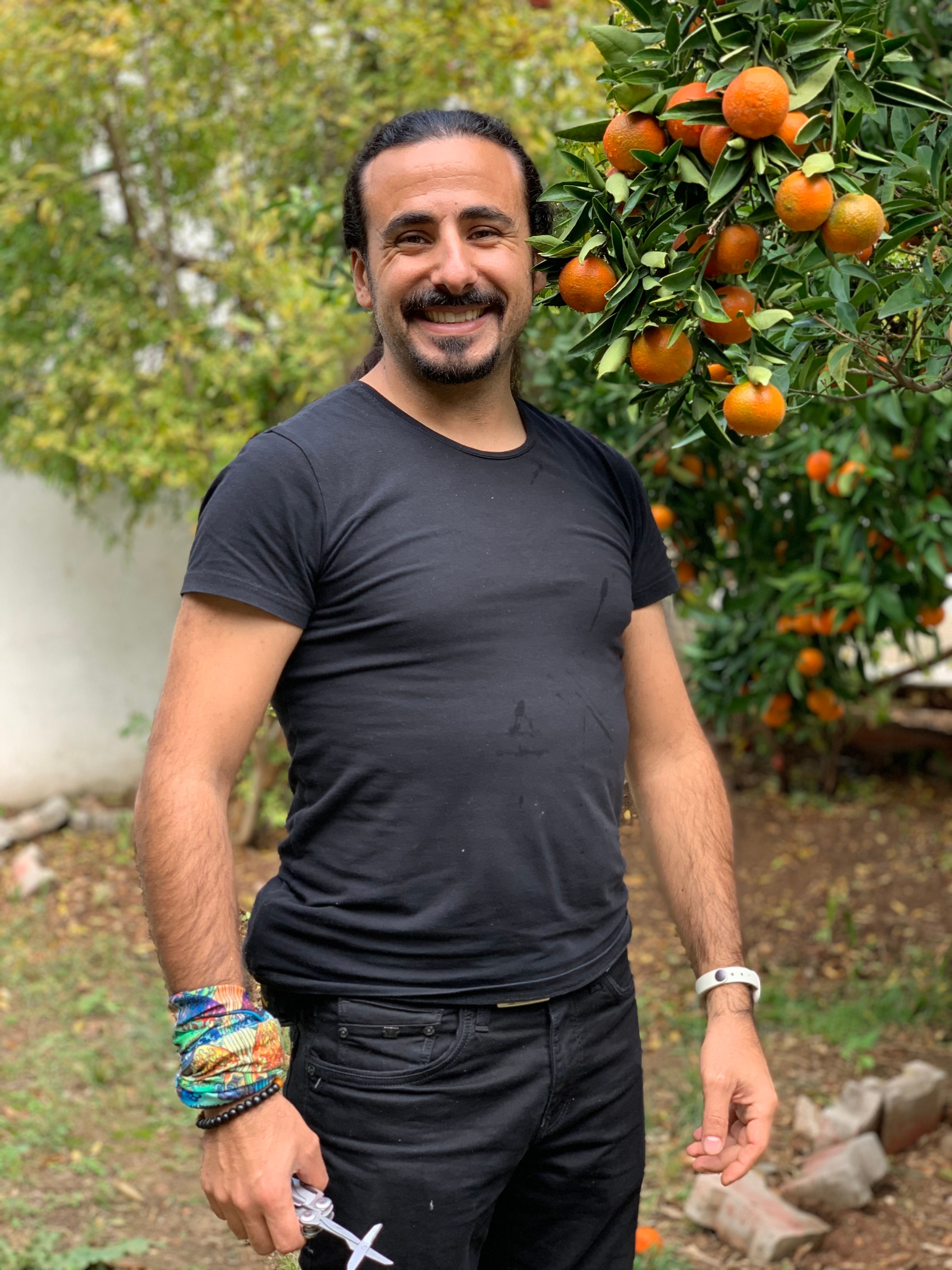 A cheerful man in black stands beside an orange tree, offering a warm and positive vibe.