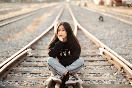 Young Girl in Black Long Sleeve Shirt Sitting on Rail Track